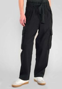 Zwarte cargobroek met een tailleband en strik, twee voorzakken en zijvakjes; gecombineerd met witte sneakers en een donker shirt.