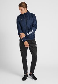 Marineblå vindjakke med høj krave, lynlås og elastiske manchetter; har hvide chevron-detaljer på ærmerne. Kombinationen er med sorte joggers og sneakers.