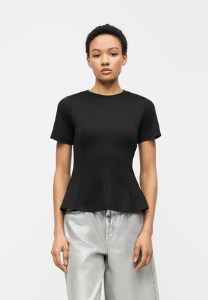 Giovane donna in piedi di fronte, indossa un top nero aderente a maniche corte e pantaloni larghi di colore argento metallico su uno sfondo semplice.