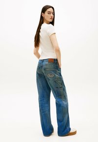 Niebieskie dżinsowe spodnie o szerokich nogawkach z widocznymi szwami kieszeni i brązową skórzaną naszywką na plecach. Połączone z dopasowaną jasną bluzką.