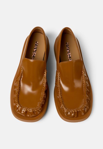 Un par de mocasines de cuero marrón, tipo slip-on, con detalles fruncidos alrededor de la punta, suelas cuadradas y plantillas de marca sobre un fondo blanco.