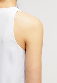 Světle šedý top bez rukávů s hladkou texturou. Má kulatý výstřih a střih, který zvýrazňuje ramena a horní část zad.