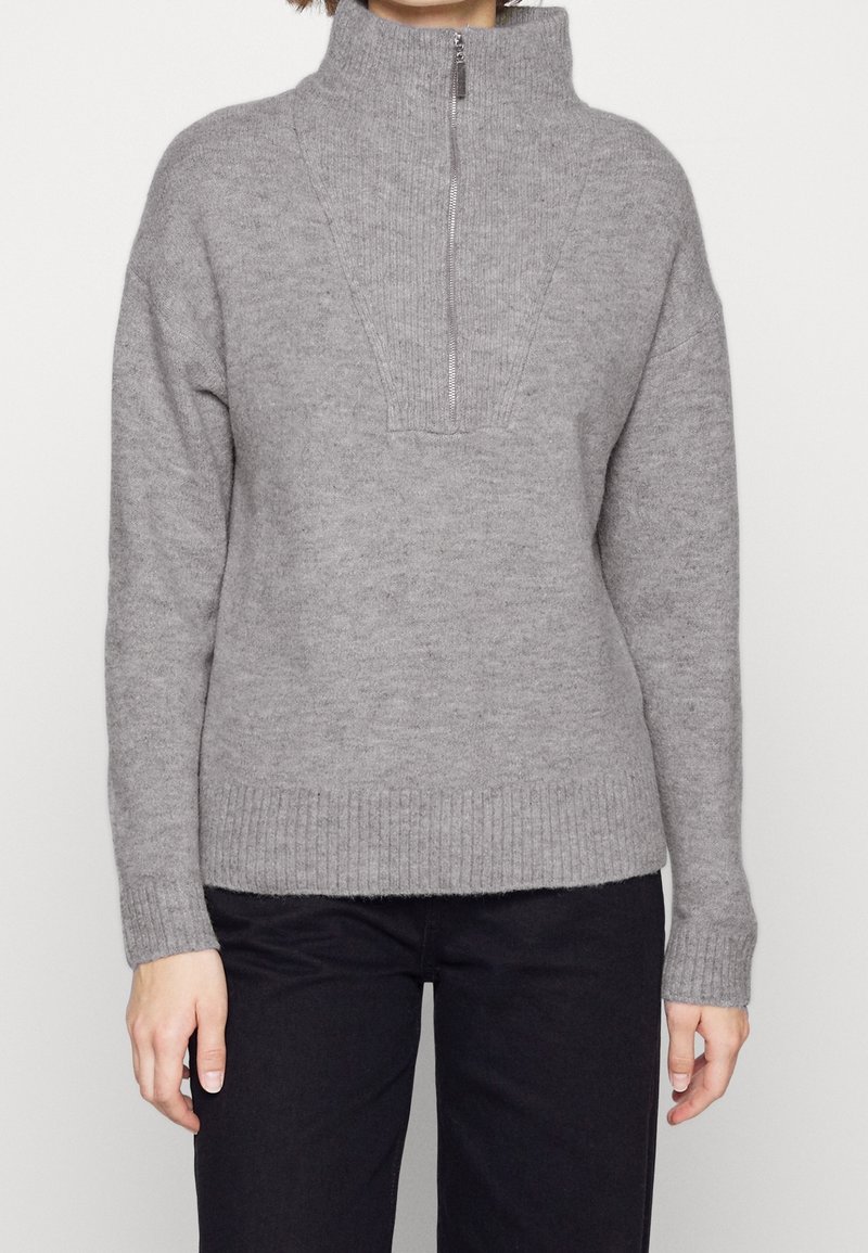 Femme portant un pull en tricot gris clair avec une fermeture éclair quart et un pantalon noir, se tenant devant un fond uni.