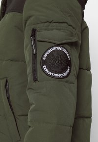 Manica di giacca verde imbottita con tasca con zip e patch rotondo nero con la scritta "Superdry® Everest Expedition Company."