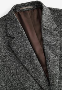 Grå tweedblazer med revers og en glat brun foer, der fremhæver et tekstureret stof med intrikate mønstre.