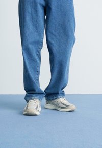 Denim plave oversized traperice s opuštenim krojem, uparene s svijetlosivim tenisicama za trčanje koje imaju bijele naglaske i teksturirane potplate.