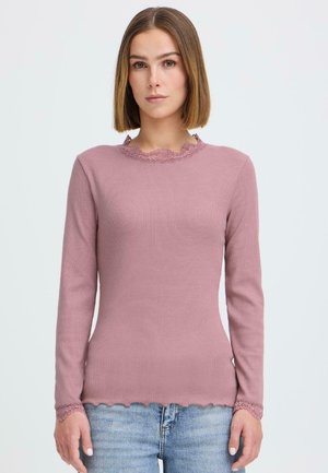 Sirgete õlgadeni juustega naine kannab lilla varrukatega pikka pluusi pitsilise äärega, kombineeritud helesiniste teksadega, seistes valge tausta ees.