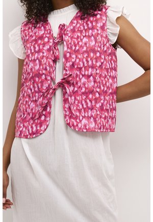 Woman wearing a white dress with ruffled sleeves and a sleeveless pink patterned quilted vest tied with two bows at the front.