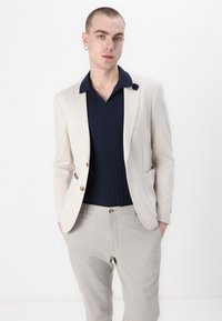 Lys beige blazer over en marineblå ribbestrikket polo, parret med lysegrå bukser. Har en knappåpning og en lomme på blazeren.