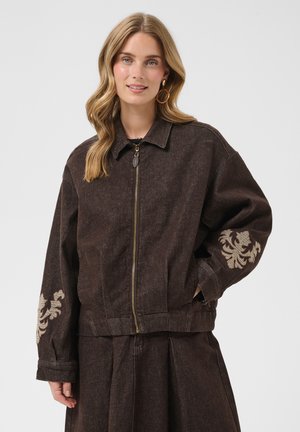 Junge Frau mit langen Haaren trägt eine dunkelbraune Reißverschlussjacke mit beiger Blumenstickerei an den Ärmeln und passenden weiten Hosen.