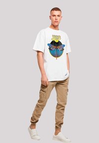 Weißes T-Shirt mit grafischem Druck, das ein mehrfarbiges Libellenmotiv zeigt, kombiniert mit beigen Cargo-Hosen und weißen Sneakern. Die Cargo-Hosen haben seitliche Taschen.
