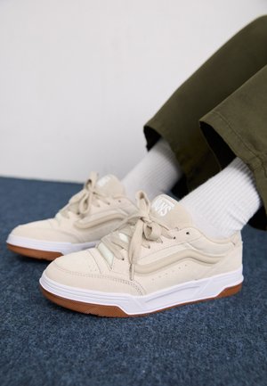 Beżowe zamszowe sneakersy z jasnooliwkowymi akcentami, sznurowane, z białymi podeszwami i teksturowaną gumową podeszwą, noszone z białymi prążkowanymi skarpetkami.