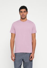 Šviesiai violetinė medvilninė marškinėliai su apvalia iškirpte ir trumpomis rankovėmis, ant krūtinės subtiliai matomas logotipas. Dėvimi su mėlynais džinsais.