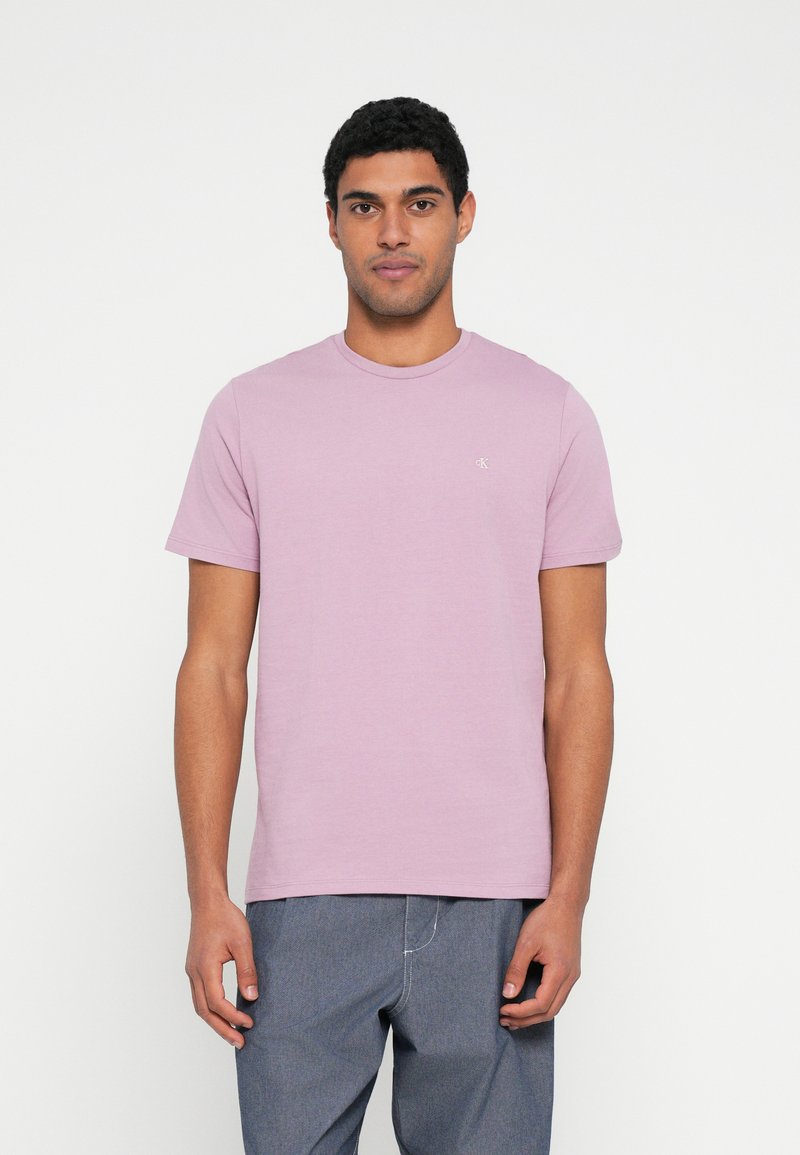 Šviesiai violetinė medvilninė marškinėliai su apvalia iškirpte ir trumpomis rankovėmis, ant krūtinės subtiliai matomas logotipas. Dėvimi su mėlynais džinsais.