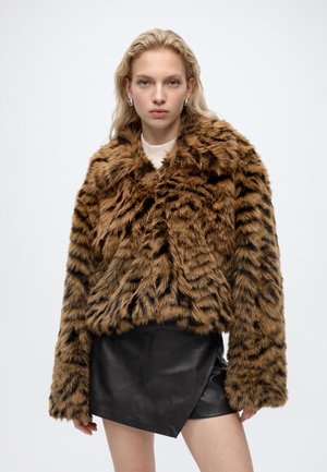 Jeune femme aux cheveux blond clair porte une veste en fausse fourrure marron et noire sur une jupe en cuir noire devant un fond uni.