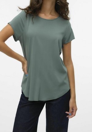 Kvinde iført en løs salviegrøn kortærmet top og mørkeblå jeans, med den ene hånd på hoften og lyseblå neglelak synlig.