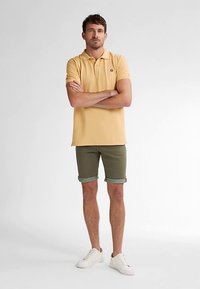 Poloshirt i lysegul med et lille logo, kombineret med olivengrønne shorts, der er rullet op i kanten, og hvide sneakers. Enkelt design og pasform.