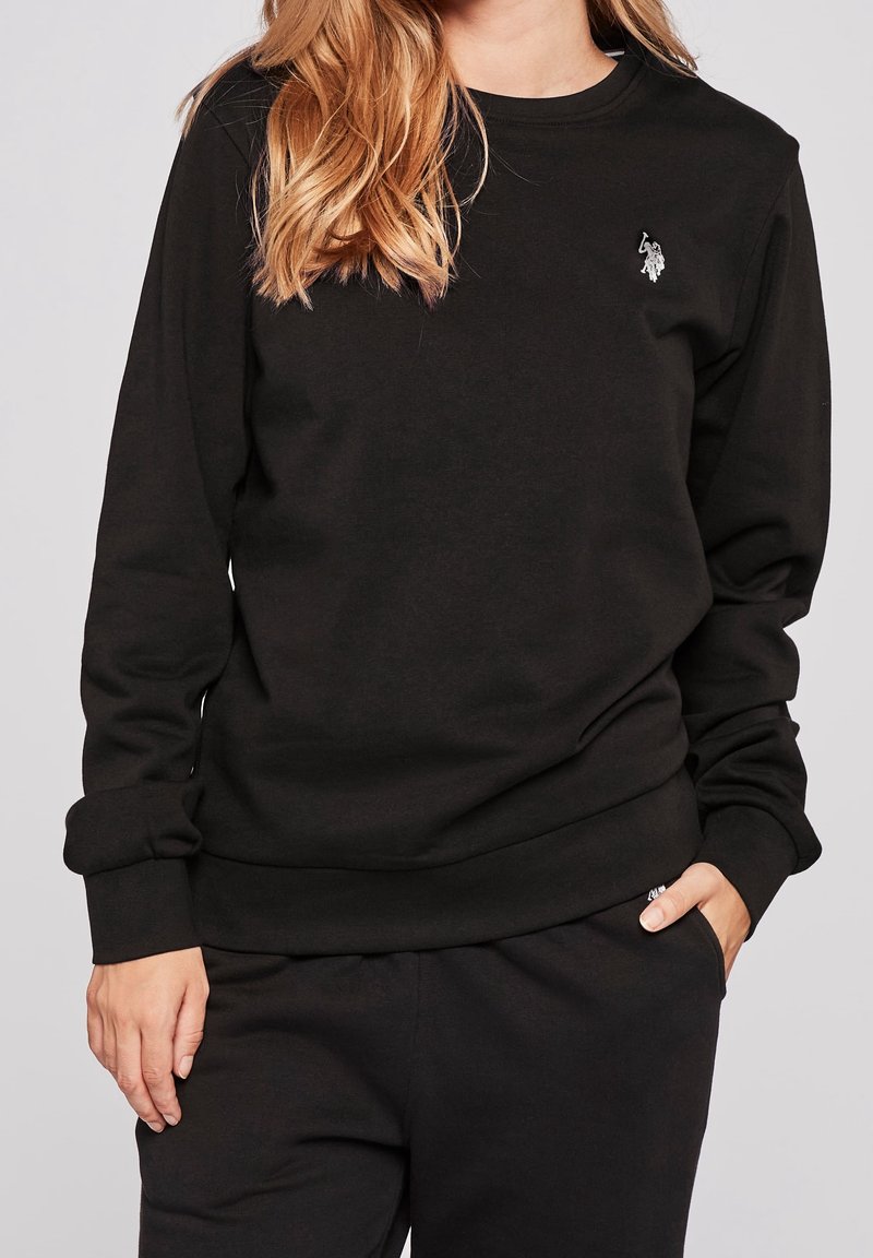 Femme portant un sweat-shirt noir à manches longues et un pantalon noir, une main dans la poche, cheveux châtains clairs reposant sur l'épaule.