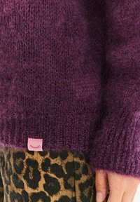 Fialový pletený sveter s huňatou textúrou, s rebrovaným lemom a malou ružovou značkou. Kombinovaný s leopardími nohavicami.