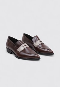 Mocasines de cuero marrón con punta afilada, que cuentan con una correa de color claro en contraste y un bajo tacón cuadrado. Textura lisa y diseño elegante.