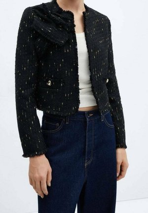 Personne portant une veste courte texturée noire avec des taches dorées, un débardeur blanc côtelé et un jean bleu foncé taille haute.