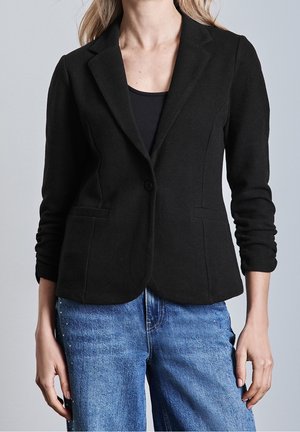Kvinde iført en tætsiddende sort blazer over en sort top og blå jeans med nitter i siderne, stående foran en ensfarvet baggrund.