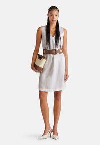 Weißes Leinenkleid ohne Ärmel mit V-Ausschnitt, braunem breiten Gürtel und beiger Handtasche; Frau trägt beige High Heels.