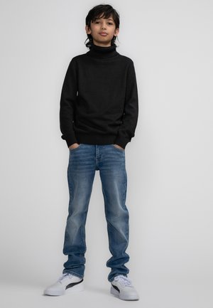 Maglione a collo alto nero, jeans blu sbiaditi e sneakers bianche. L'outfit presenta un design aderente, un tessuto morbido e una vestibilità rilassata.