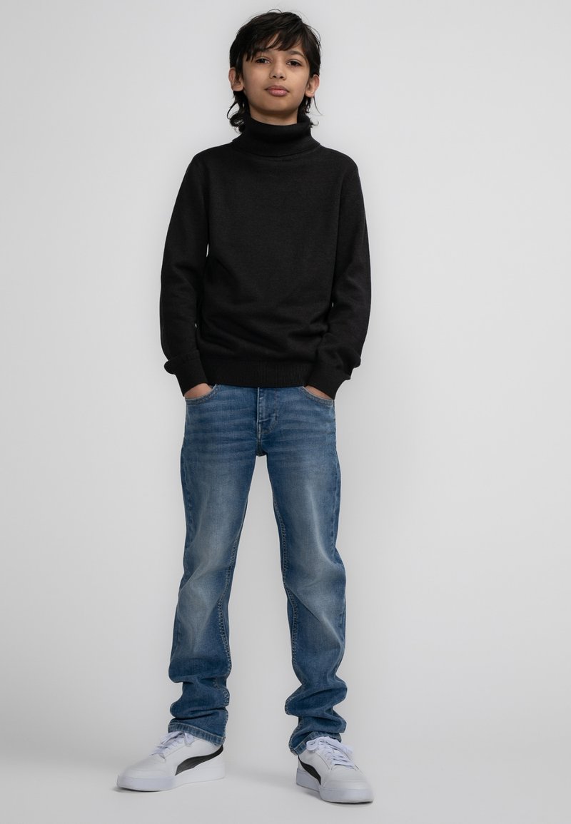 Maglione a collo alto nero, jeans blu sbiaditi e sneakers bianche. L'outfit presenta un design aderente, un tessuto morbido e una vestibilità rilassata.