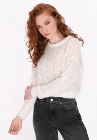Weißer, leichter Strickpullover mit strukturiertem Muster und gerippten Bündchen, kombiniert mit hochgeschnittenen schwarzen Jeans.