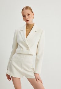 Witte tweed cropped jacket met dubbele parelknopen, gecombineerd met bijpassende high-waisted shorts. Gladde textuur, scherpe revers en gestructureerd ontwerp.