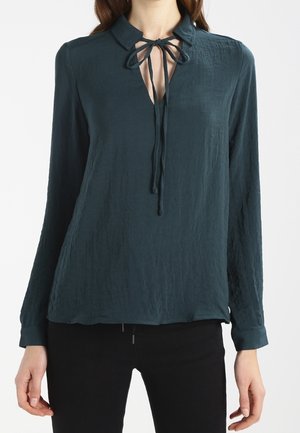 Femme portant un chemisier à manches longues bleu sarcelle foncé avec un col et un détail de nœud à l’avant, associé à un pantalon noir.