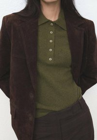 Blazer de ante marrón oscuro sobre un polo de punto verde oliva con cuello y tapeta de cinco botones. Tela texturizada con pantalones de rayas verticales.