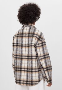 Kostkovaná flanelová košile v krémové, šedé a rezavé barvě. Má rozepínací límec, dlouhé rukávy a volný střih s zapínáním na knoflíky.