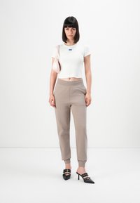 Jonge vrouw staat tegen een witte achtergrond in een witte cropped T-shirt, taupe joggerpants, zwarte hoge muilen en draagt een lichtroze schoudertas.