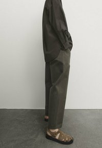 Dunkelbraunes Hemd mit Knopf, lockere olivgrüne Hose und braune Sandalen mit Riemenverzierungen und schwarzer Sohle, vor einem neutralen Hintergrund.