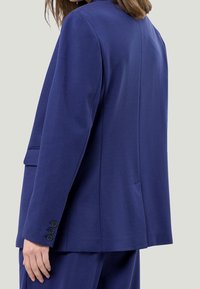 Blazer bleu marine à coupe décontractée, doté de revers crantés et de poignets à quatre boutons. Tissu lisse avec une texture minimale et une fente au dos.