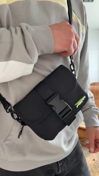 Musta kankaiset olkalaukku, jossa on läppäkiinnitys, säädettävä hihna ja muovinen solki. Edessä logo korostettuna vihreänä.