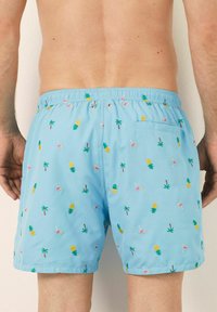 Shorts de baie albastru deschis cu modele brodate de ananas, palmieri și cocktail-uri. Buzunar elastic la talie și un singur buzunar la spate.