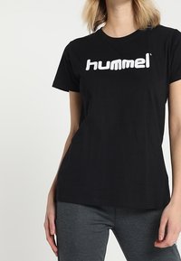 Camiseta de algodón negra con un destacado logo blanco de "hummel", cuello redondo, manga corta y un corte relajado. Diseño casual con detalles mínimos.