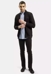 Czarna kurtka na zamek błyskawiczny noszona na jasnoniebieskiej koszuli z guzikami, w komplecie z czarnymi spodniami i czarnymi butami. Gładka tkanina, swobodny sportowy krój.