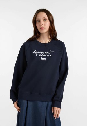 Giovane donna con capelli lunghi e castani che indossa una felpa blu navy con testo bianco in corsivo e una piccola illustrazione di un cane, abbinata a una gonna blu.