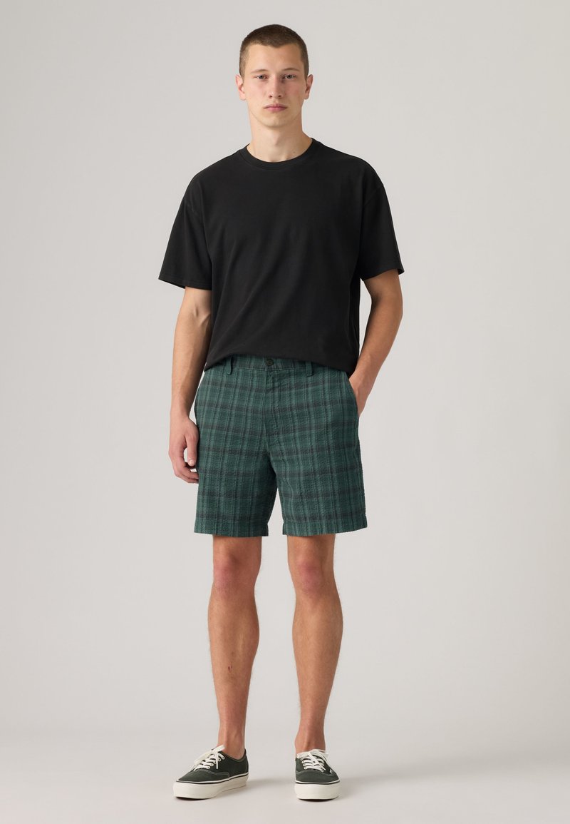 Giovane uomo in piedi con indosso una t-shirt nera, pantaloncini verdi a quadri e sneakers verdi con suola bianca su sfondo semplice.