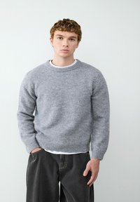 Grijze wollen trui met een ronde hals, lange mouwen en geribbelde boorden. Gecombineerd met zwarte wijde broek en een witte shirt eronder.