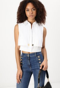 Camicetta bianca senza maniche, corta, con pieghe e dettagli in oro, abbinata a jeans blu a vita alta con bottoni dorati. Borsa nera in mano.