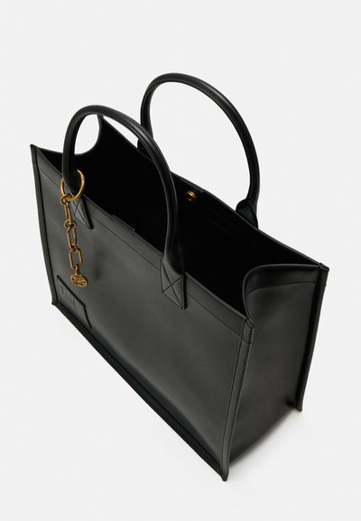 Sac en cuir noir avec double poignées, chaîne dorée avec médaillon et fermeture à bouton pression, présenté légèrement ouvert sur fond blanc.