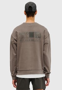 Brun sweatshirt av bomull, med stort tryck på baksidan som inkluderar text och mönster, samt muddade ärmslut och fåll.