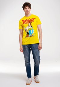 Gelbes T-Shirt mit einem grafischen Druck von Asterix und Obelix. Kurze Ärmel, Rundhals, aus Baumwollmaterial. Tragen zu blauen Jeans und Sneakers.