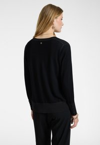 Maglione nero a maglia con maniche lunghe, scollatura circolare e dettagli con bottoni sui polsini. Presenta un logo sulla nuca e una texture morbida.