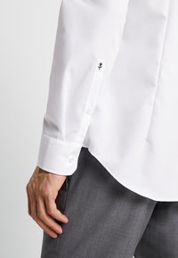 Witte button-up blouse van een gladde stof, met een zwarte rozenborduursel op de manchetten en afgeronde zoom, geaccessorieerd met grijze broek.